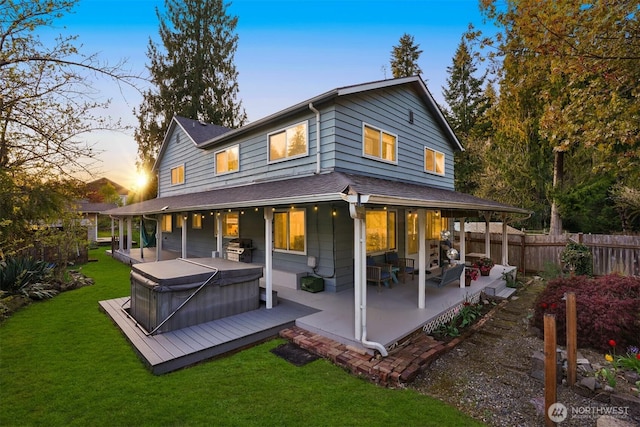back of property at dusk with a lawn, a shingled roof, fence, a hot tub, and a patio