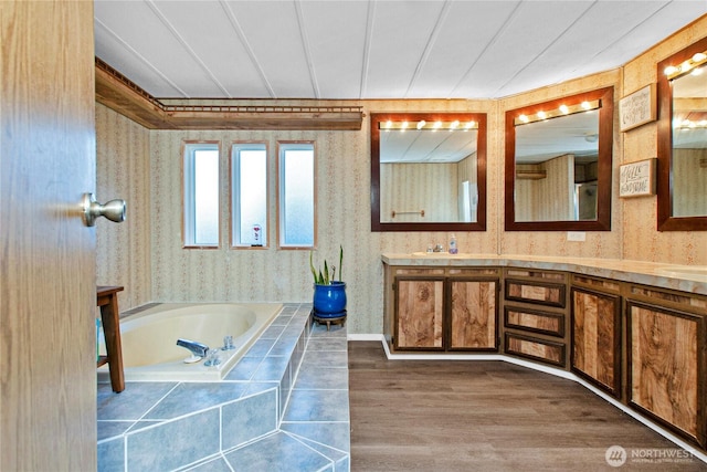 full bathroom featuring double vanity, wallpapered walls, a bath, and wood finished floors
