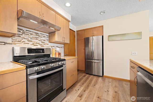 kitchen featuring appliances with stainless steel finishes, under cabinet range hood, light wood-type flooring, light countertops, and decorative backsplash