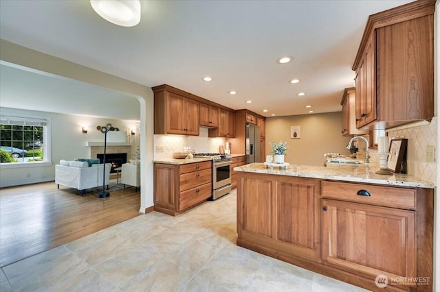 kitchen with stainless steel appliances, a lit fireplace, a peninsula, brown cabinetry, and light stone countertops