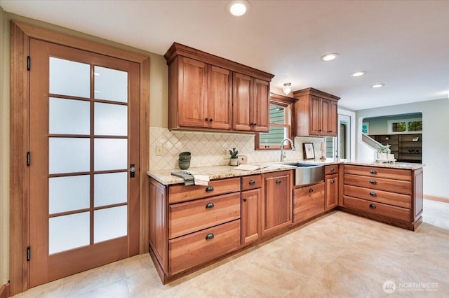 kitchen with a sink, light stone counters, decorative backsplash, brown cabinets, and a peninsula