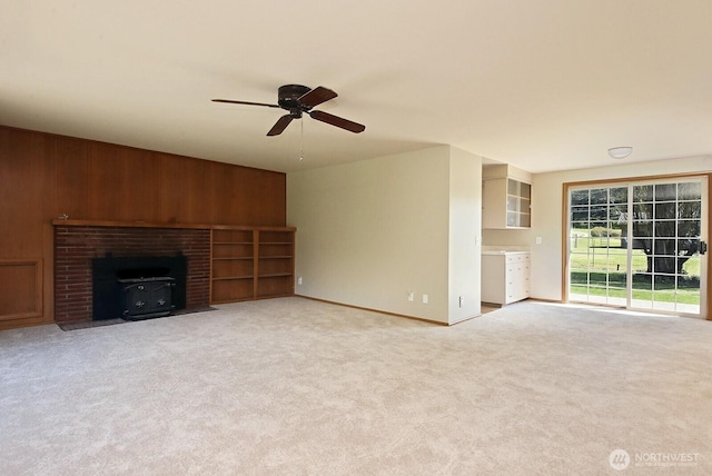 unfurnished living room featuring baseboards, light colored carpet, wood walls, ceiling fan, and a wood stove