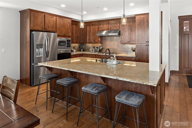 kitchen featuring appliances with stainless steel finishes, a sink, tasteful backsplash, under cabinet range hood, and dark wood-type flooring