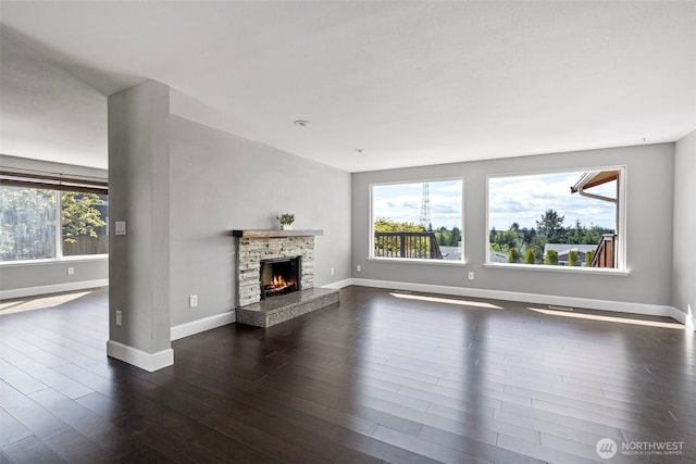 unfurnished living room featuring a stone fireplace, baseboards, a wealth of natural light, and dark wood finished floors