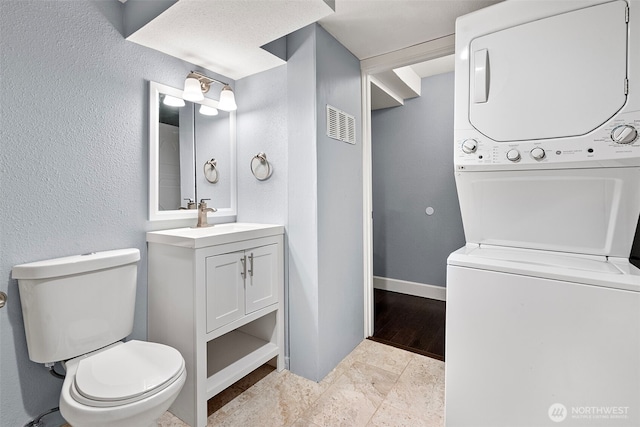 bathroom featuring toilet, baseboards, stacked washer / drying machine, a textured wall, and vanity