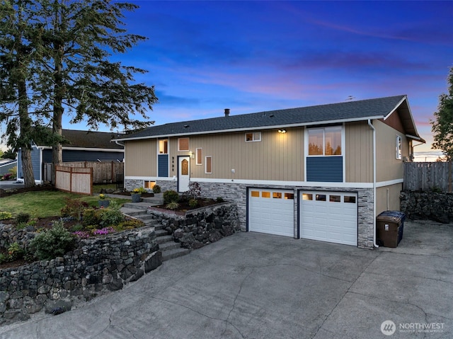 view of front of house featuring roof with shingles, driveway, stone siding, fence, and a garage