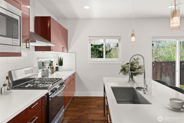 kitchen with light countertops, wall chimney range hood, a sink, appliances with stainless steel finishes, and dark wood-style flooring