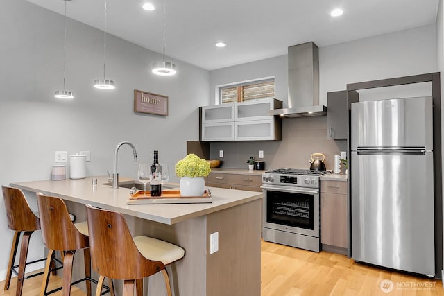 kitchen with appliances with stainless steel finishes, wall chimney range hood, a kitchen bar, light wood finished floors, and a sink