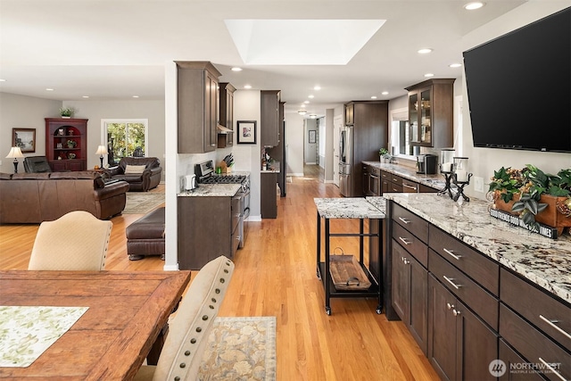 kitchen with stainless steel appliances, open floor plan, glass insert cabinets, light wood finished floors, and dark brown cabinetry
