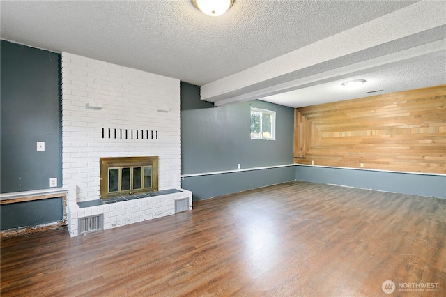 unfurnished living room featuring a brick fireplace, a textured ceiling, wood finished floors, wooden walls, and beam ceiling