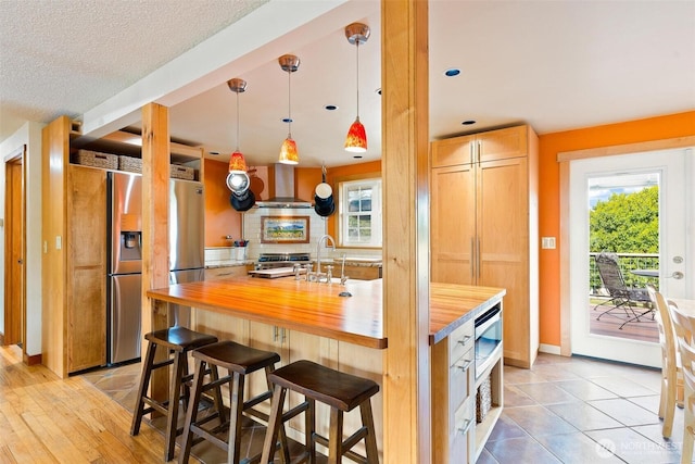 kitchen with wooden counters, wall chimney exhaust hood, appliances with stainless steel finishes, tasteful backsplash, and a kitchen island with sink