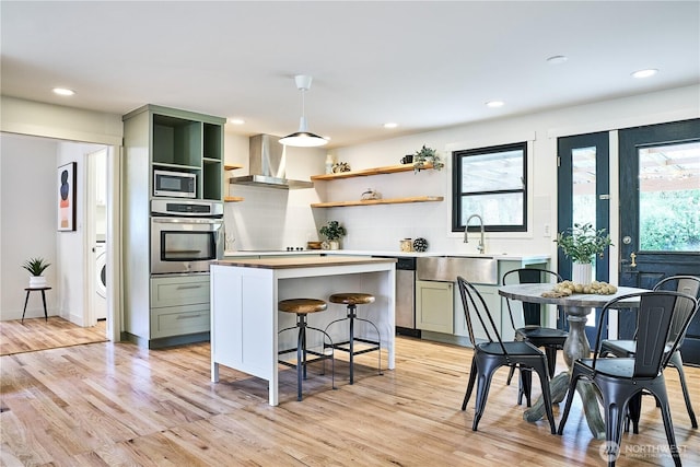 kitchen featuring appliances with stainless steel finishes, wall chimney range hood, open shelves, light wood-type flooring, and a kitchen island