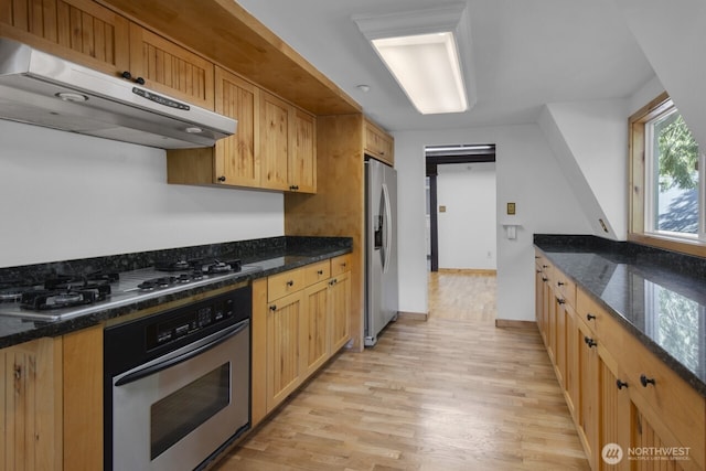 kitchen with under cabinet range hood, appliances with stainless steel finishes, dark stone counters, and light wood-style flooring