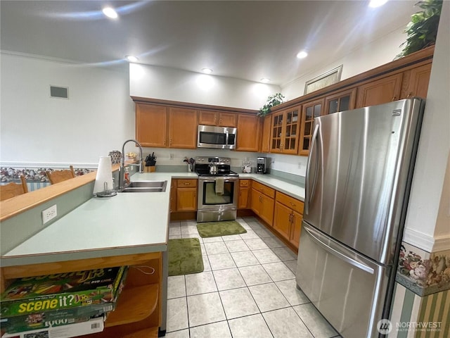 kitchen with glass insert cabinets, brown cabinetry, light tile patterned floors, a sink, and stainless steel appliances