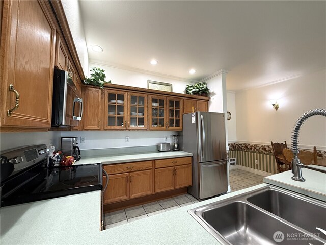 kitchen with stainless steel appliances, brown cabinetry, a sink, crown molding, and glass insert cabinets