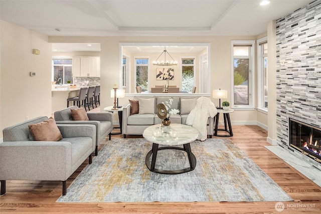 living area with a raised ceiling, wood finished floors, a stone fireplace, baseboards, and a chandelier