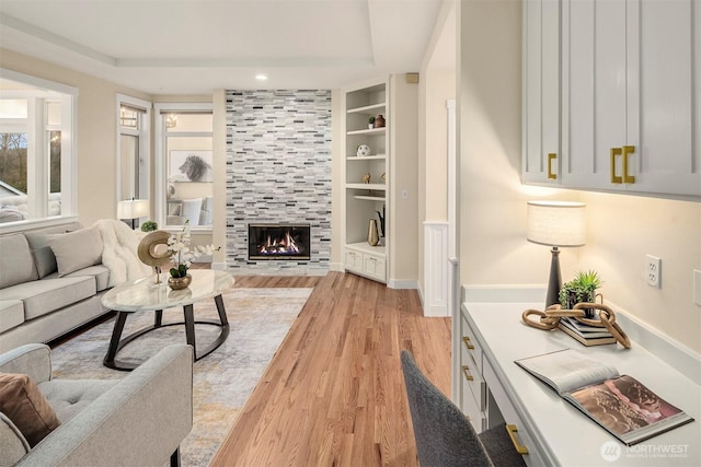 living room with light wood-type flooring, built in features, a fireplace, and a tray ceiling