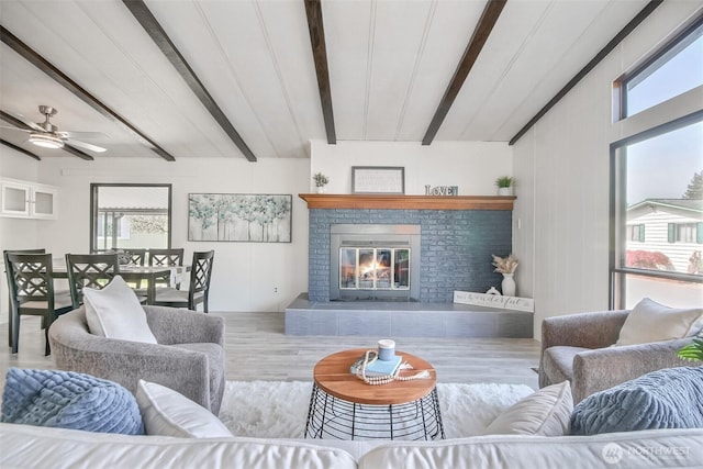 living room with a brick fireplace, ceiling fan, beam ceiling, and wood finished floors