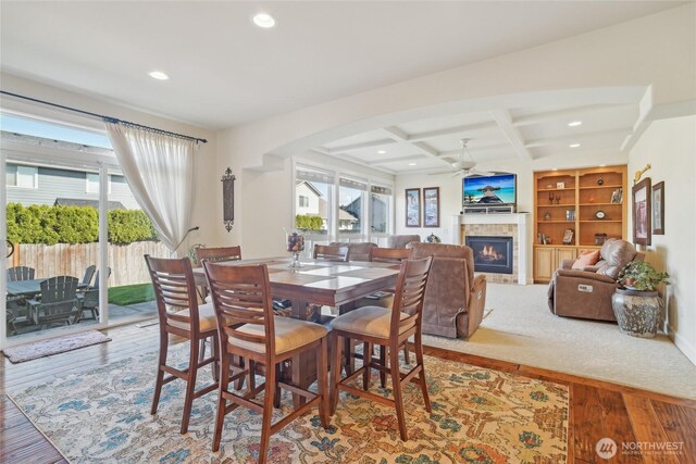 dining area with a tiled fireplace, wood finished floors, coffered ceiling, beamed ceiling, and ceiling fan