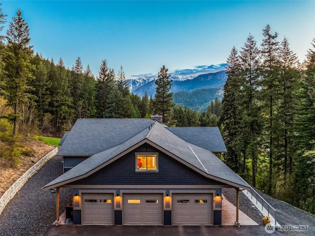 view of front facade featuring a garage, roof with shingles, a chimney, a mountain view, and a wooded view