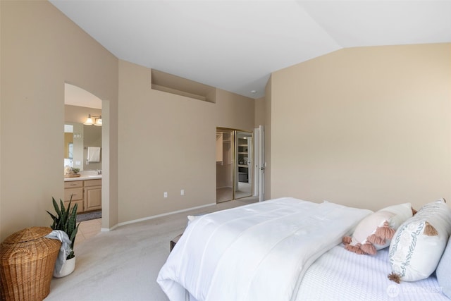bedroom featuring vaulted ceiling, light colored carpet, ensuite bath, baseboards, and arched walkways