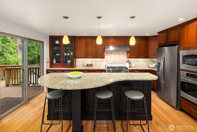 kitchen featuring stainless steel appliances, under cabinet range hood, light wood-type flooring, tasteful backsplash, and a kitchen bar