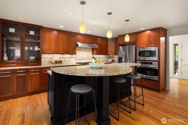kitchen featuring stainless steel appliances, under cabinet range hood, light wood-style floors, backsplash, and a kitchen bar