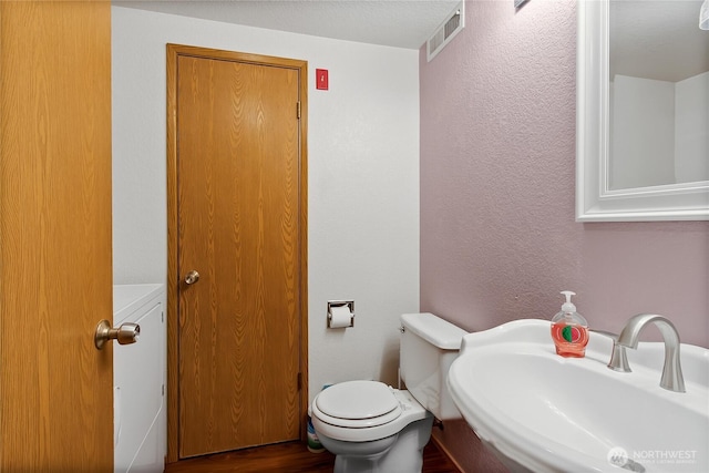 half bath with wood finished floors, a sink, a textured wall, toilet, and visible vents