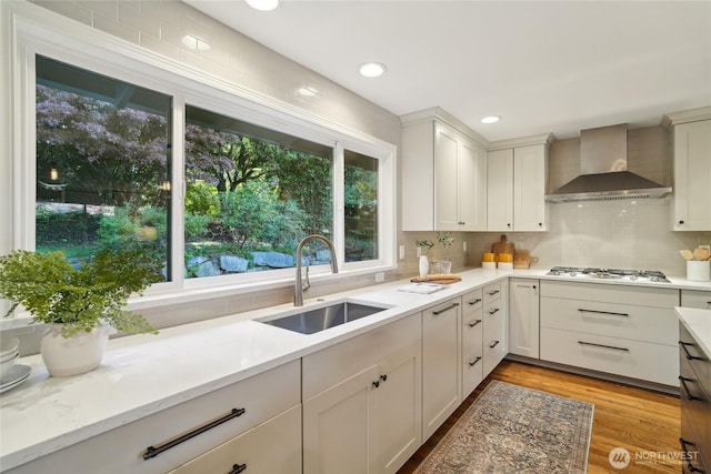 kitchen with wall chimney range hood, a sink, light wood finished floors, stainless steel gas stovetop, and backsplash