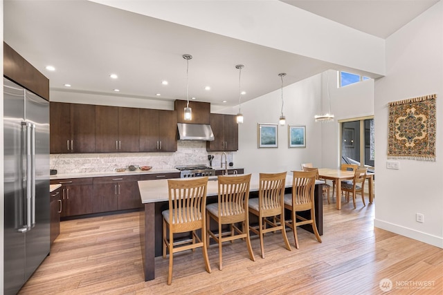 kitchen with dark brown cabinetry, light countertops, backsplash, under cabinet range hood, and stainless steel built in refrigerator