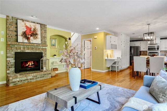 living room featuring a brick fireplace, light wood-type flooring, baseboards, an inviting chandelier, and ornamental molding
