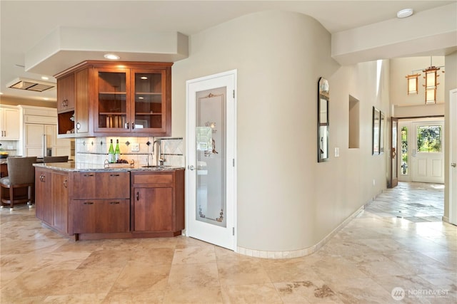 kitchen featuring brown cabinetry, tasteful backsplash, a sink, stone countertops, and glass insert cabinets