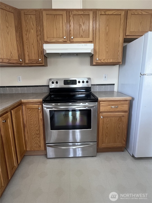 kitchen featuring under cabinet range hood, electric range, brown cabinetry, and freestanding refrigerator