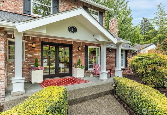 entrance to property with a shingled roof, brick siding, a chimney, french doors, and covered porch