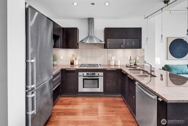 kitchen with stainless steel appliances, a sink, wall chimney exhaust hood, a peninsula, and light stone countertops