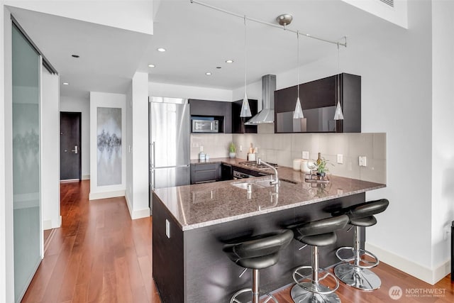 kitchen with appliances with stainless steel finishes, wall chimney range hood, a peninsula, light stone counters, and baseboards