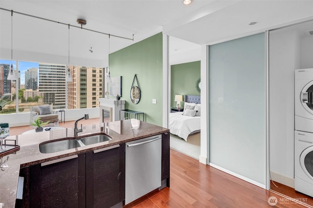 kitchen featuring dishwasher, a sink, stacked washer and clothes dryer, light wood-style flooring, and dark stone countertops