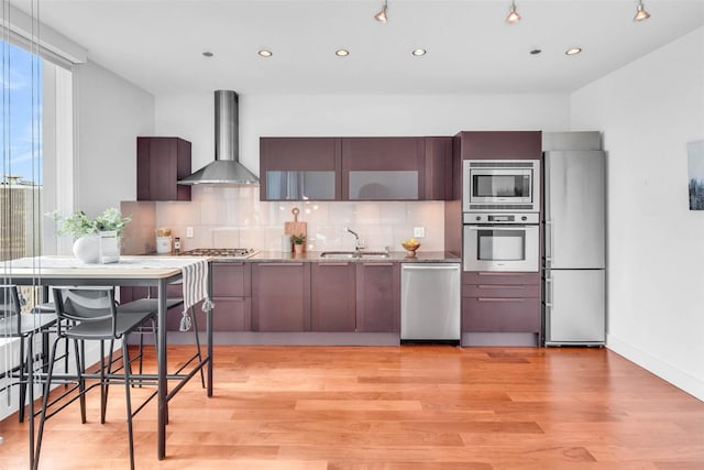 kitchen featuring appliances with stainless steel finishes, wall chimney range hood, a sink, decorative backsplash, and light wood-type flooring