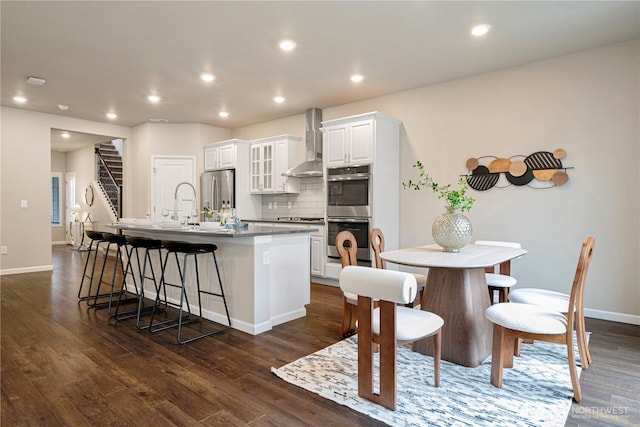 kitchen with wall chimney range hood, a kitchen island with sink, a kitchen breakfast bar, stainless steel appliances, and tasteful backsplash