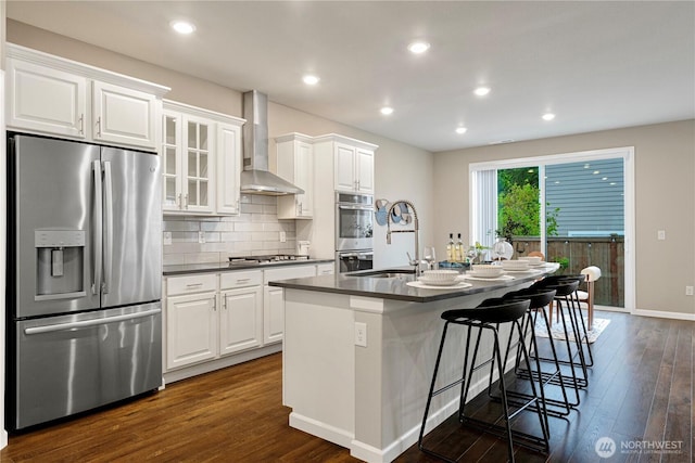 kitchen with wall chimney range hood, dark wood-style flooring, dark countertops, appliances with stainless steel finishes, and a center island with sink