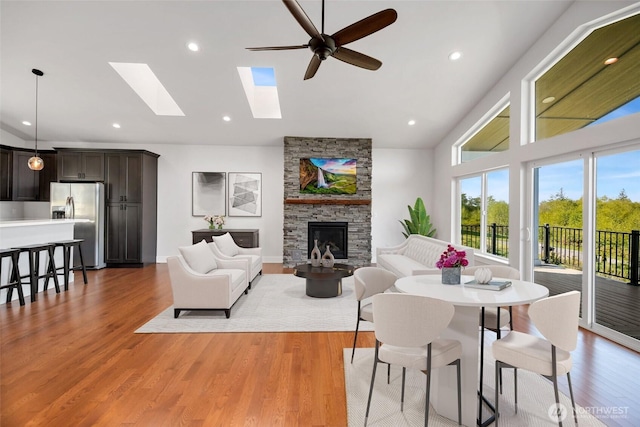 living room featuring a stone fireplace, ceiling fan, high vaulted ceiling, light wood-type flooring, and a skylight