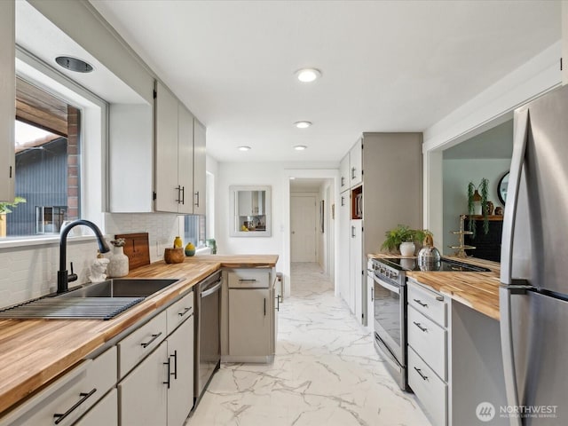 kitchen featuring wooden counters, appliances with stainless steel finishes, a sink, light marble finish flooring, and tasteful backsplash