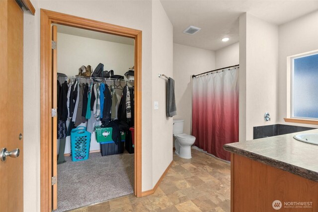 bathroom with visible vents, toilet, vanity, a walk in closet, and baseboards