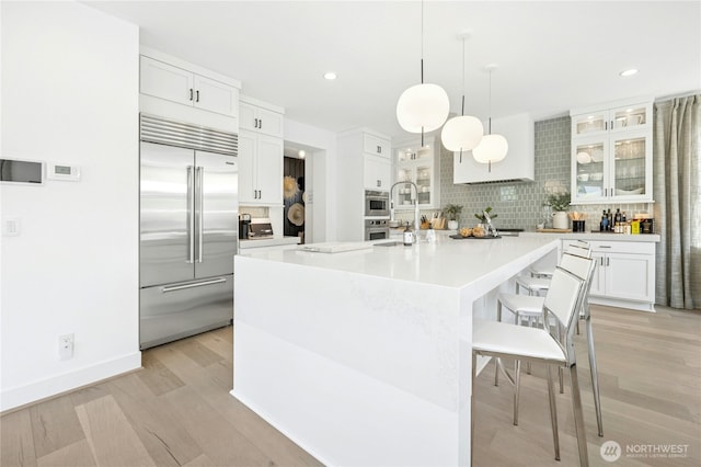 kitchen featuring a breakfast bar area, stainless steel built in refrigerator, light wood-style flooring, glass insert cabinets, and backsplash