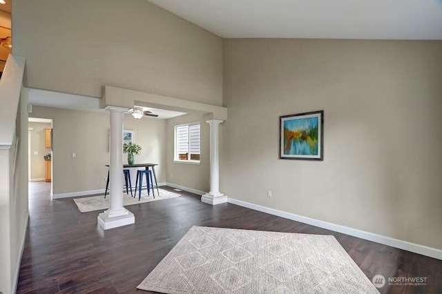 empty room featuring ceiling fan, a high ceiling, baseboards, ornate columns, and dark wood-style floors