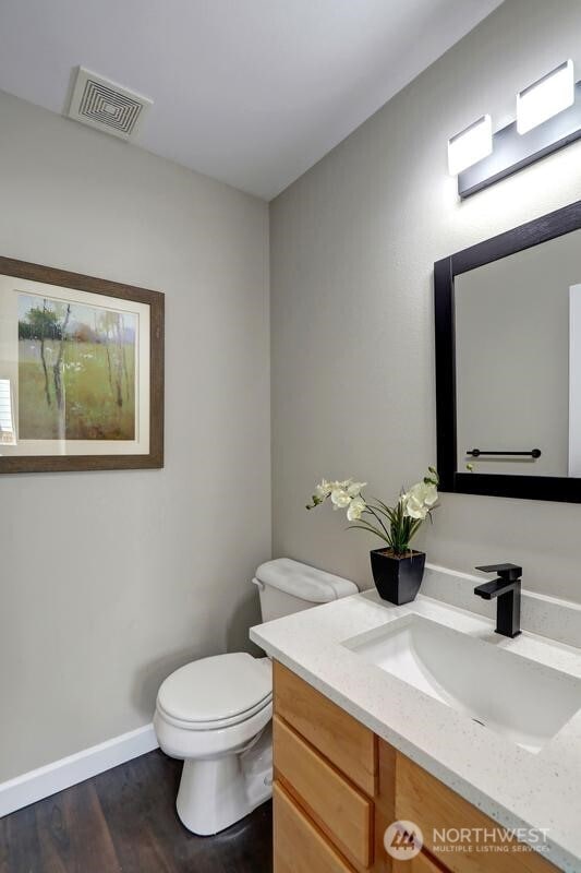 half bath with visible vents, wood finished floors, vanity, baseboards, and toilet