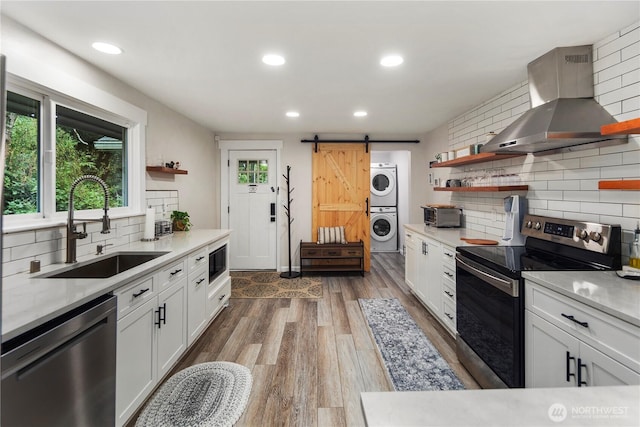 kitchen with open shelves, stacked washer / dryer, wall chimney range hood, a sink, and appliances with stainless steel finishes