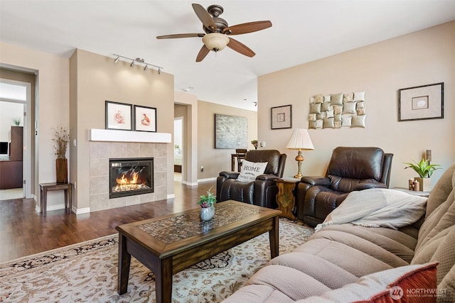 living room featuring baseboards, rail lighting, wood finished floors, a tiled fireplace, and a ceiling fan
