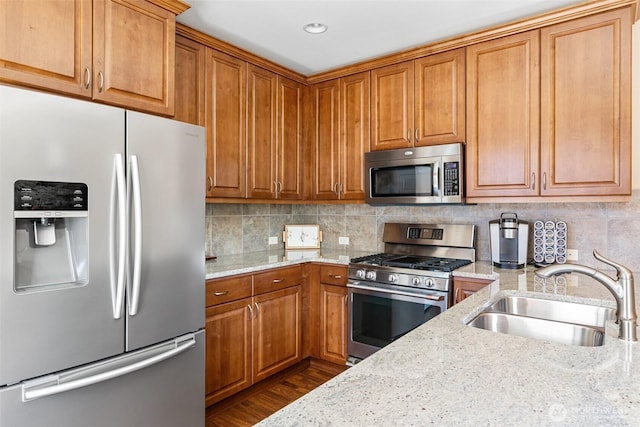 kitchen with light stone countertops, decorative backsplash, brown cabinetry, appliances with stainless steel finishes, and a sink