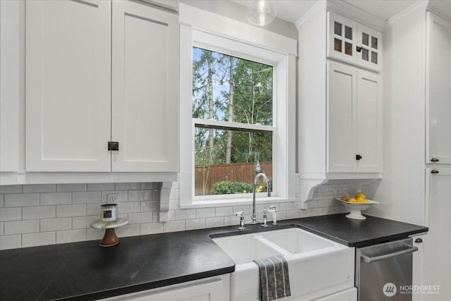 kitchen featuring stainless steel dishwasher, dark countertops, a sink, white cabinets, and backsplash